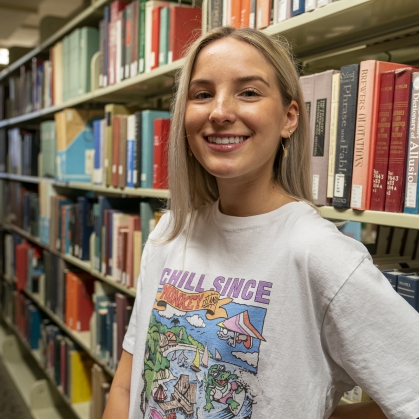 a female college student standing in front of shelves of books in the library